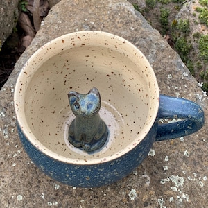 Könnte beinhalten: Eine blaue Keramiktasse mit gesprenkeltem beigefarbenem Innenraum, mit einer kleinen blauen Katzenfigur im Inneren. Die Tasse hat einen blauen Griff und steht auf einer Steinoberfläche. Die Maße der Tasse sind in Zentimetern.