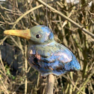 Könnte beinhalten: Ein blauer und grüner Keramikvogel mit gelbem Schnabel und schwarzen Augen. Der Vogel sitzt auf einem braunen Ast.