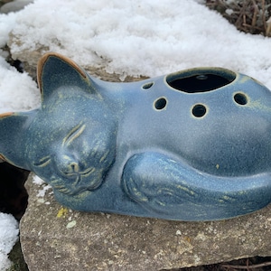 Könnte beinhalten: Eine blaue Keramik-Katzenfigur mit einem schlafenden Gesicht und einem Loch im Rücken zum Pflanzen. Die Katze liegt auf der Seite mit den Pfoten unter dem Körper.