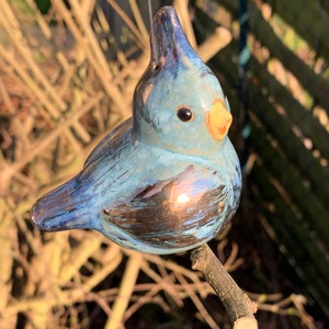 Könnte beinhalten: Ein blauer Keramikvogel-Anhänger mit goldener Metallic-Oberfläche. Der Vogel hat einen gelben Schnabel und schwarze Augen. Er hängt an einem Zweig.