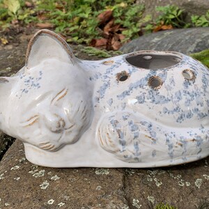 Könnte beinhalten: Ein keramischer Katzen-Blumentopf in Schlafposition. Der weiße Topf hat blaue, gesprenkelte Akzente und braune Verzierungen. Er hat drei runde Löcher zum Bepflanzen. Die Augen der Katze sind geschlossen und ihre Ohren sind aufgestellt.