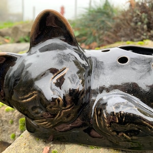 Könnte beinhalten: Eine schwarze Keramik-Katzenfigur mit glänzender Oberfläche. Die Katze liegt mit geschlossenen Augen und hat ein kleines Loch im Rücken zum Pflanzen.