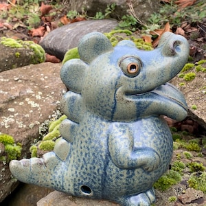 Könnte beinhalten: Eine keramische Drachenskulptur in Blau- und Grüntönen. Die Figur hat eine strukturierte Oberfläche und ein cartoonartiges Design. Sie befindet sich auf einem moosbewachsenen Felsen und schafft so eine natürliche Umgebung. Die Skulptur ist ein dekorativer Gegenstand.
