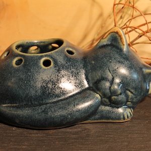 Könnte beinhalten: Eine blaue Keramik-Katzenfigur mit einem schlafenden Gesicht. Die Katze hat einen ausgehöhlten Rücken mit Löchern zum Aufbewahren kleiner Gegenstände.