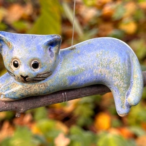 Könnte beinhalten: Eine blaue Keramik-Katzenfigur mit gesprenkeltem Glasur, die auf einem Ast liegt. Die Katze hat schwarze Augen und eine schwarze Nase.