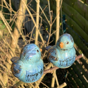 Könnte beinhalten: Zwei keramische Blaumeisen-Ornamente mit glänzendem Finish sitzen auf einem kleinen Ast. Die Vögel haben braune Akzente und sind an dünnen Drähten aufgehängt. Der Hintergrund zeigt eine verschwommene Ansicht eines Holzzauns und von Ästen.