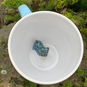 Könnte beinhalten: Weiße Keramiktasse mit hellblauem Henkel. Im Inneren der Tasse befindet sich eine kleine, türkis- und bronzefarbene Keramikfigur. Die Tasse steht auf einer moosigen Oberfläche.
