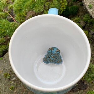 Könnte beinhalten: Weiße Keramiktasse mit hellblauem Henkel und einer türkis-braunen Froschfigur im Inneren. Die Tasse hat ein türkisfarbenes Band um den Boden. Die Tasse steht auf einem moosigen Felsen.