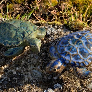 Könnte beinhalten: Zwei Keramik-Schildkrötenfiguren. Eine ist türkis mit dunkelblauen Akzenten, die andere blau mit braunen Akzenten. Beide haben strukturierte Panzer und werden auf einer Steinoberfläche präsentiert. Dekorationsartikel.