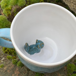 Könnte beinhalten: Weißer Keramikbecher mit hellblauem Henkel und Rand. Im Inneren der Tasse befindet sich eine kleine, türkisblaue Katzenfigur. Die Tasse steht auf einem Bett aus grünem Moos.