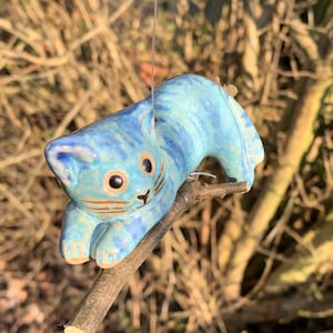 Könnte beinhalten: Eine blaue Keramik-Katzenfigur mit schwarzen Augen und Schnurrhaaren, die auf einem Ast sitzt. Die Katze ist mit einem hellblauen und weißen Marmormuster bemalt.