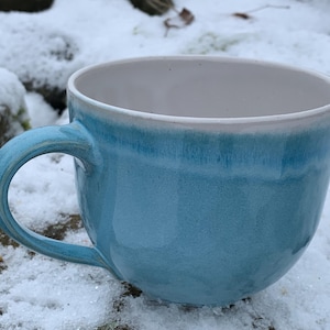 Könnte beinhalten: Eine Keramiktasse mit hellblauem Äußeren und weißem Innenraum. Die Tasse hat eine abgerundete Form mit einem großen Henkel. Die Tasse steht auf einer schneebedeckten Oberfläche.
