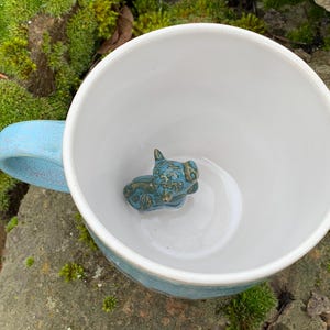 Könnte beinhalten: Eine weiße Keramiktasse mit hellblauem Henkel und Rand. Im Inneren der Tasse befindet sich eine kleine, türkisfarbene Keramik-Tierfigur. Die Tasse steht auf einem moosigen Felsen.