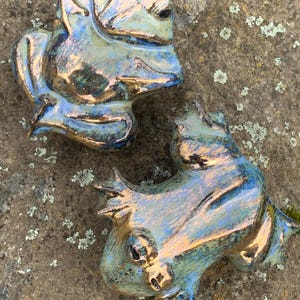 Könnte beinhalten: Zwei Keramik-Froschfiguren mit einem blauen und grünen irisierenden Überzug. Die Frösche sitzen auf einer strukturierten braunen Oberfläche.
