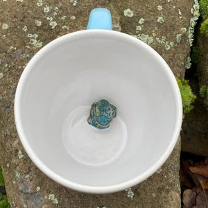 Könnte beinhalten: Weiße Keramiktasse mit hellblauem Henkel. Am Boden der Tasse befindet sich eine kleine, türkisgrüne Froschfigur. Die Tasse steht auf einer moosigen, strukturierten Felsfläche.
