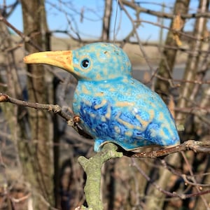 Könnte beinhalten: Eine blaue und beige Keramikvogel-Figur mit gelbem Schnabel, die auf einem Ast sitzt. Der Vogel hat einen Draht zum Aufhängen. Der Hintergrund ist verschwommen und zeigt Äste und einen Hauch von Himmel. Geeignet für den Innen- und Außenbereich.