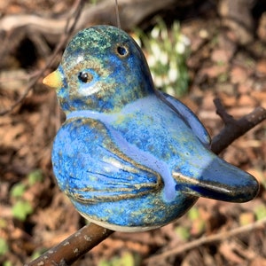 Könnte beinhalten: Ein blauer und grüner Keramikvogel-Anhänger mit gelbem Schnabel. Der Vogel hängt an einer Schnur und sitzt auf einem Ast.