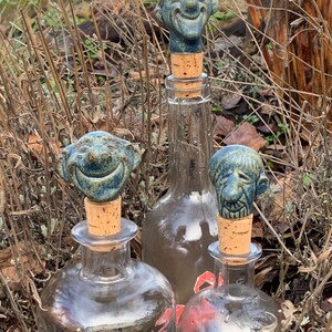 Könnte beinhalten: Drei Glasflaschen mit dekorativen Stöpseln. Jeder Stöpsel hat ein blaues Keramikgesicht mit einem lächelnden Ausdruck. Die Flaschen sind klar und die Stöpsel sind aus Kork. Die Flaschen sind von getrockneten Pflanzen umgeben.