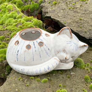 Könnte beinhalten: Weiße Keramik-Katzenfigur mit blauen und braunen Akzenten. Die Katze liegt in einer schlafenden Position zusammengerollt mit geschlossenen Augen. Die Figur hat eine hohle Mitte mit Löchern zum Halten von Blumen.