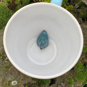 Könnte beinhalten: Eine weiße Keramiktasse mit hellblauem Henkel und einem teal-farbenen, vogelförmigen Design im Boden der Tasse. Die Tasse steht auf einer moosigen Oberfläche.