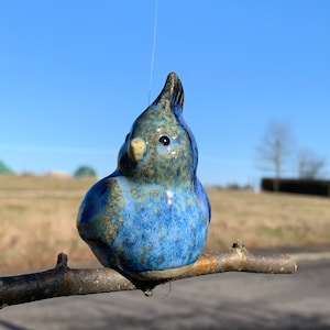 Könnte beinhalten: Eine blaue und grüne Keramik-Vogelfigur, die auf einem Ast sitzt. Der Vogel hat eine hellblaue Glasur mit dunkelblauen Flecken und einen hellgrünen Schnabel.