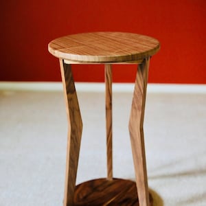 May include: A handcrafted wooden side table with a round top and a unique geometric leg design. The table is made of light and dark brown wood, with a circular base. The table is approximately 20 inches tall.