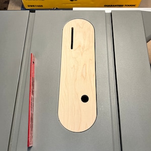 May include: A close-up of a gray table saw with a wooden insert. The insert has a slot and a round hole. The table saw has a red ruler on the left side.