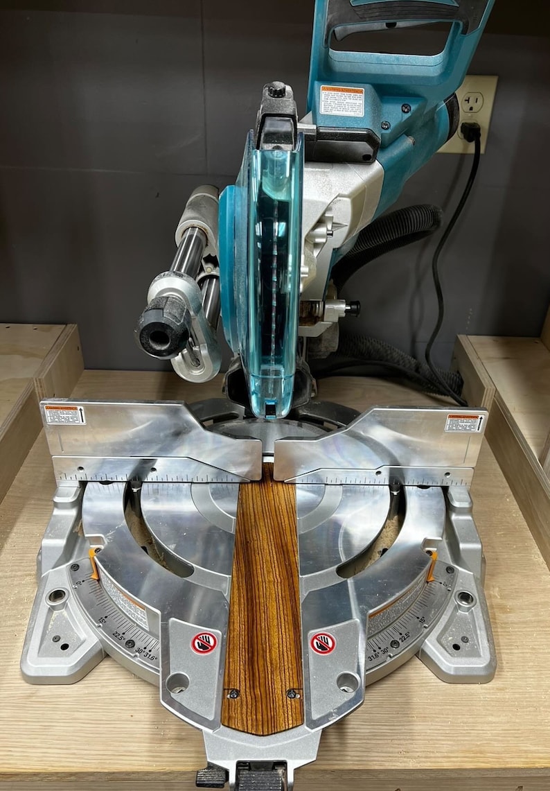 K&ouml;nnte beinhalten: Nahaufnahme einer silbernen Gehrungss&auml;ge mit einem Holzbrett, das zum Schneiden positioniert ist. Die S&auml;ge ist auf einen Winkel von 45 Grad eingestellt und hat einen blauen und silbernen S&auml;geblattschutz.