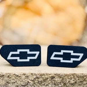 May include: Two black rectangular emblems with a white Chevrolet bow tie logo. The emblems are displayed on a stone surface with a blurred background. The Chevrolet logo is a recognizable symbol of the automotive brand.