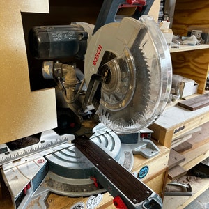 May include: A gray and black Bosch miter saw with a clear plastic blade guard. The saw is on a wooden table with a brown wooden board on the table. The saw has a black and brown handle and a red lever. There are several stickers on the saw, including one that says "The Masters".