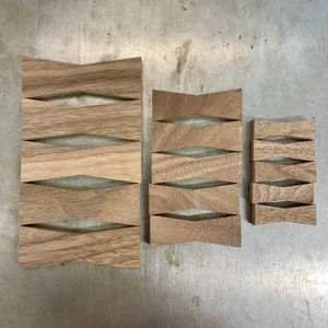 Puede incluir: Tres paneles decorativos de madera de diferentes tamaños. Cada panel está hecho de madera marrón claro y presenta una serie de cortes en forma de diamante. Los paneles están dispuestos sobre una superficie metálica, mostrando la veta natural de la madera.