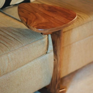 May include: A wooden side table with a rounded top and a single leg that extends from the top to the base. The table is attached to a couch and has a natural wood finish.