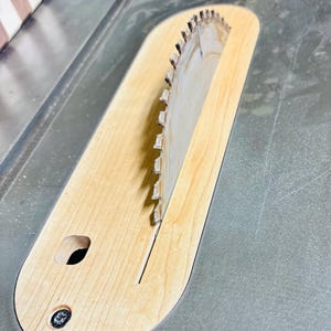 May include: A wooden table saw blade guard with a circular saw blade. The guard is made of light-coloured wood and has a curved shape. The saw blade is silver and has sharp teeth. The guard is attached to a grey table.