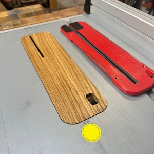 May include: A close-up of a wooden table saw fence with a red and black adjustment lever. The fence is labeled "Precision Cut - Use Soft Pencil -  T-Track Lock - Adjust to Cut Length".