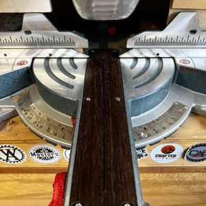 May include: A close-up view of a miter saw with a wooden cutting board in place. The saw is silver and black with a red handle. The cutting board is dark brown with a groove down the center. The saw has a protractor with markings in degrees.