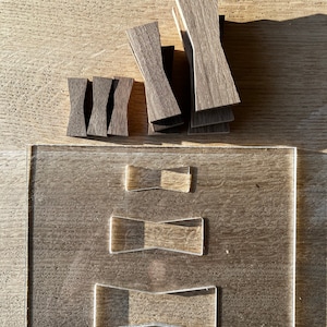 Puede incluir: Un conjunto de formas de pajarita de madera, apiladas sobre una superficie de madera, con una plantilla de acrílico transparente que muestra tres tamaños diferentes de formas de pajarita.