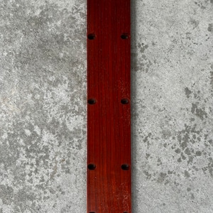 May include: A long, narrow, wooden board with a rounded top and bottom. The board has six evenly spaced holes drilled into it. The board is a reddish-brown color.