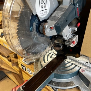 May include: A silver and black miter saw with a clear plastic guard. The saw has a black wooden fence and a red lever. The saw is labeled "DEWALT". A white sticker with the text "dd ww daniel durlap woodworks" is on the saw.