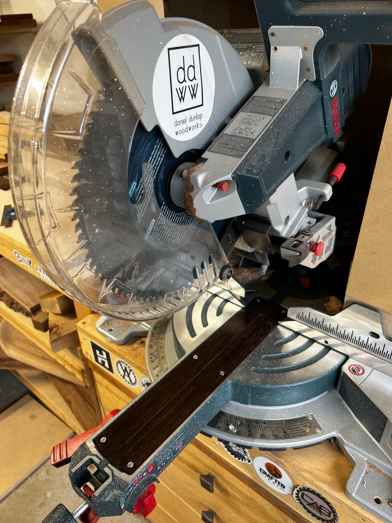 May include: A silver and black miter saw with a clear plastic guard. The saw has a black wooden fence and a red lever. The saw is labeled "DEWALT". A white sticker with the text "dd ww daniel durlap woodworks" is on the saw.