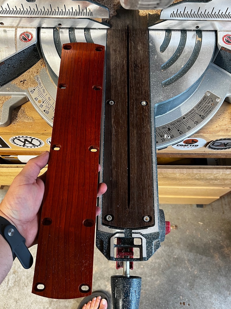 May include: A close-up of a dark brown wooden board with a slot cut into it. The board is being held by a hand wearing a black wristband. The board is being held over a silver and black miter saw.