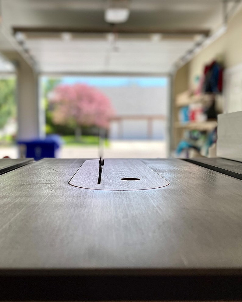 May include: A close-up of a table saw blade slot with a metal blade in place. The blade is in a recessed area of the table saw surface. The background is blurred and shows a garage with a door open to the outside.