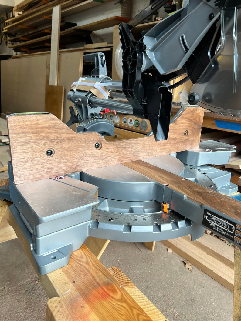 Peut inclure: Gros plan d'une scie &agrave; onglets avec une planche de bois en cours de coupe. La scie est pos&eacute;e sur une table en bois et est de couleur gris et noir. La scie a un rapporteur sur sa base pour mesurer les angles.