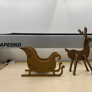 May include: A brown cardboard reindeer with a red nose and a brown cardboard sleigh. The reindeer and sleigh are on a light brown surface. The text "SHAPEOKO" is visible on a silver bar in the background.