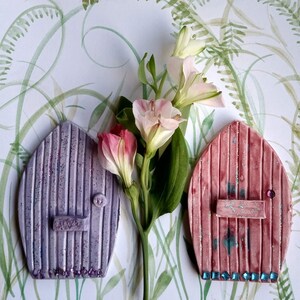 May include: Two decorative fairy doors, one purple and one pink, with glitter accents and a floral design. The doors are on a white background with green watercolor accents.
