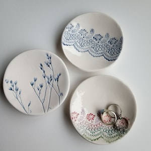 May include: Three ceramic ring dishes with different designs. One dish features a blue floral pattern, another has a blue and white lace design, and the third has a green and pink floral design with a lace border.