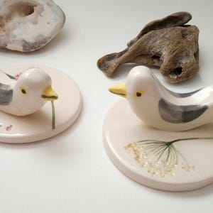 May include: Two ceramic bird ornaments. Each bird has a white and grey body, yellow beak, and black eye. They sit on a round, pale pink base. One base has the word "bird", the other a floral design. Decorative home decor.