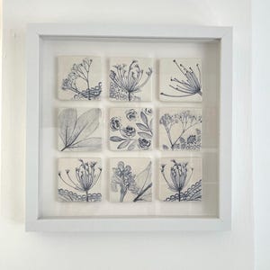 May include: A white square frame containing nine individual square ceramic tiles arranged in a three-by-three grid. Each tile features a dark blue botanical illustration, including various flowers, leaves, and lace-like patterns on a white background.