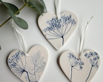 May include: Three white ceramic heart-shaped ornaments with blue pressed flower designs. Each heart has a white ribbon for hanging.