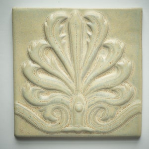 May include: A square ceramic tile with a raised, white, floral design. The tile has a light green background.