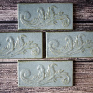 May include: Four rectangular, pale blue ceramic tiles featuring a raised, decorative floral pattern. The tiles are displayed on a wooden surface, highlighting the intricate relief details and colour variations. Each tile measures approximately 15 cm x 7.5 cm.